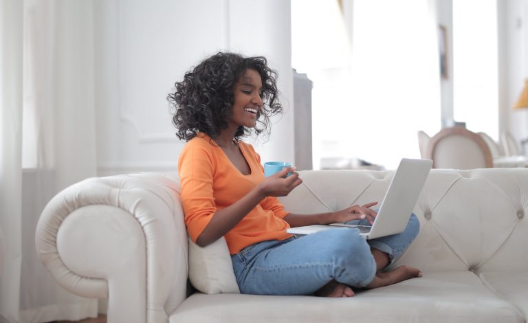 smiling woman in orange shirt on couch with laptop and coffee